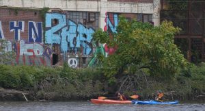 Kayakers Mary Bruno and guide Carl Alderson go past a building with graffiti situated along the Passaic River in the film, American River, a discussion of environmental justice. Photo Credit: Scott Morris Productions